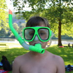 Ein Junge trägt eine knallgrüne Taucherbrille und Schnorchel. Im Hintergrund sieht man eine Liegewiese und Badesee.