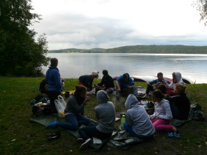 Eine Gruppe Jugendlicher sitzt auf einer Wiese. Im Hintergrund ist ein See oder Fluss zu sehen, an dessen Ufer einige Kanus liegen. Die Jugendlichen sitzen im Kreis und essen gemeinsam.