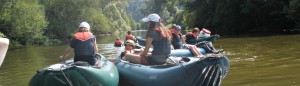 Mehrere Kanus auf einem Fluss. Die Kinder und Jugendlichen in den Kanus tragen Schwimmwesten, sie entspannen gerade und sind nicht am Paddeln.
