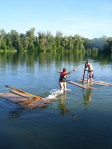 Es sind zwei Kinder mit etwas Abstand auf einem See umgeben von ein paar Holzplanken. Eines steht auf Holzplanken, das andere versucht auf den Holzplanken zu laufen. Es schaut aus, als ob ein selbstgebasteltes Floß auseinander gefallen ist und die beiden Kinder nun mit den Resten des Floßes spielen.