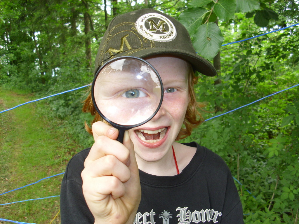 Ein lachender Junge mit Schildmütze hält ein Vergrößerungsglas vor sein Gesicht und schaut hindurch. Durch das Vergrößerungsglas ist sein eines Auge stark vergrößert.
