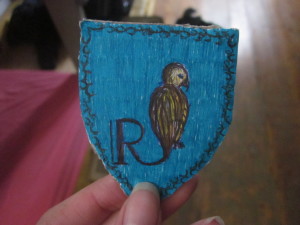 Ein selbstgebasteltes Wappen der Ravenclaws. Ein sitzender Vogel auf blauem Hintergrund mit dem "R" für Ravenclaw.
