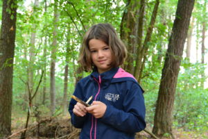 Ein Mädchen steht im Wald und hält zwei Zweige in der Hand, so als ob sie den einen mit dem anderen schnitzen möchte.