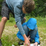 Ein Junge kniet auf einer Wiese am Boden und hat verschiedene Bestimmungsbücher aufgeschlagen. Ein Betreuer hilft dem Jungen bei der Bestimmung verschiedener Pflanzen.